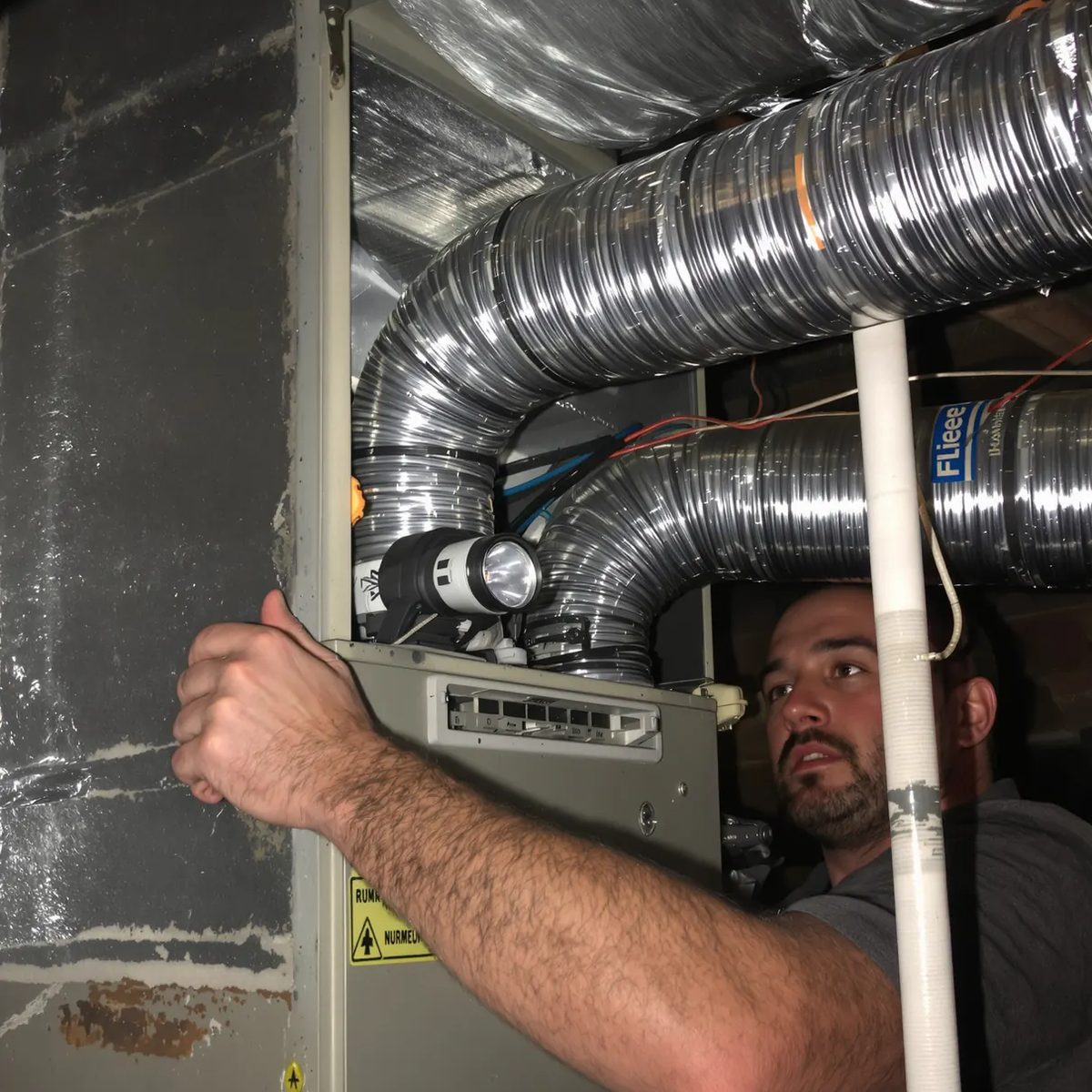 An HVAC technician reaching into an air handler surrounded by silver flex duct