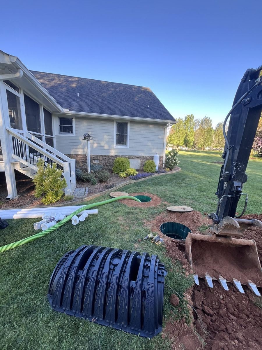 A new black plastic septic tank on the lawn beside an open dig, with an excavator bucket and a house in the background