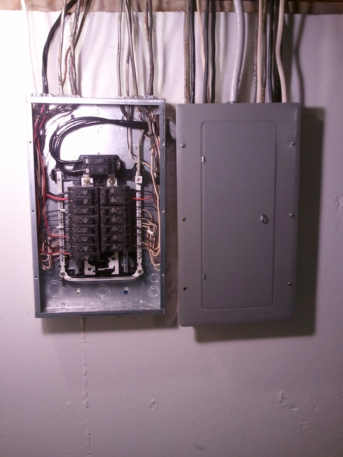 A home breaker panel and a subpanel on a basement wall, with the cover off one panel and many cables entering from the top