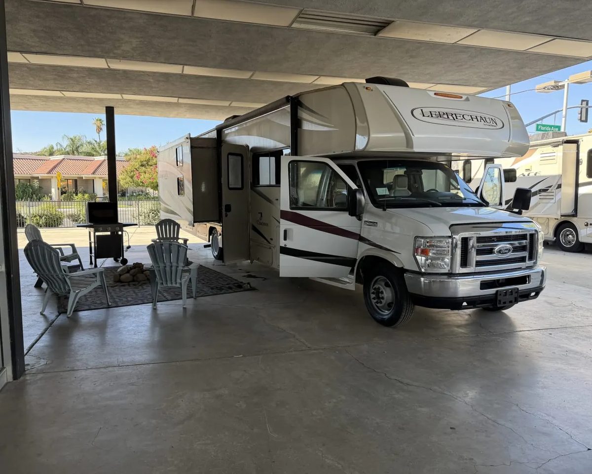 A Coachmen Leprechaun Class C motorhome under a covered lot with its awning out and chairs set up for a renter