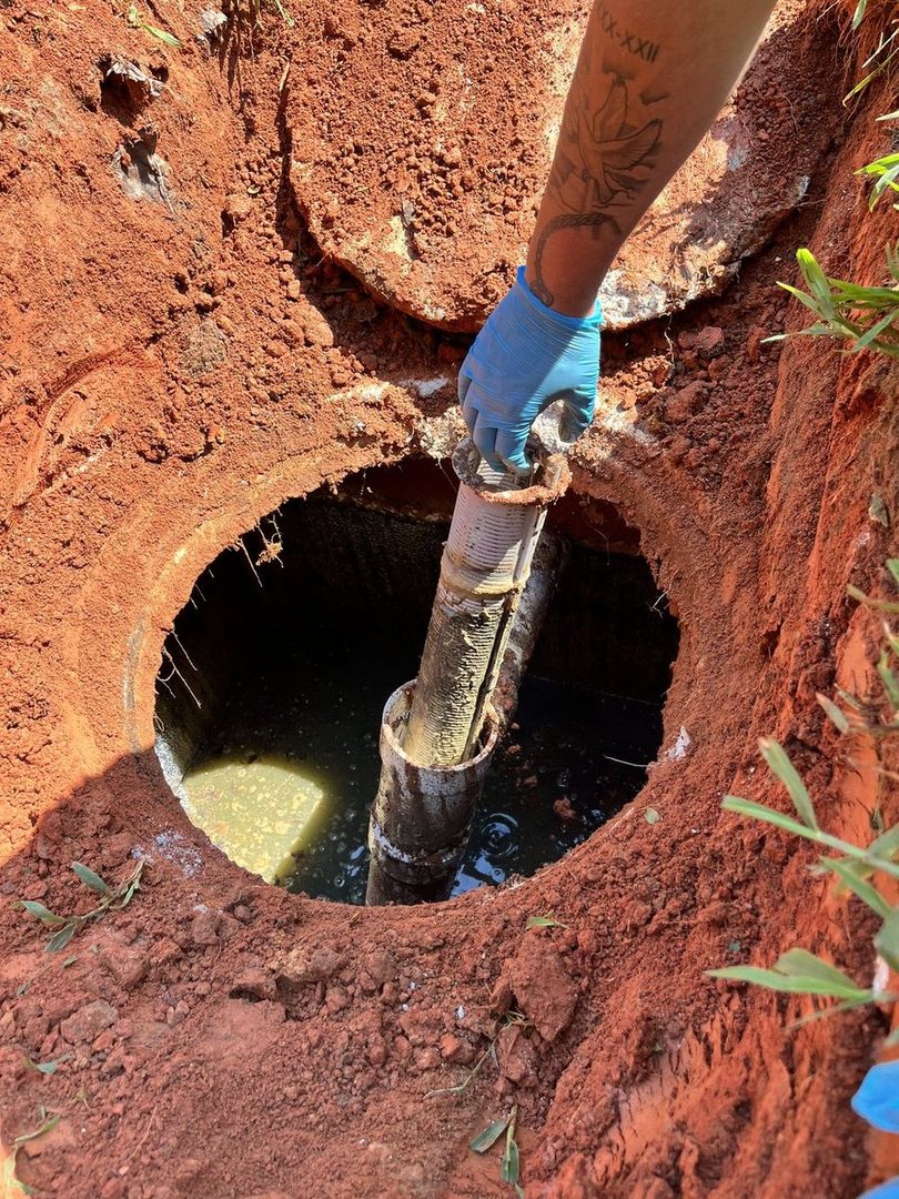 A septic tech's gloved hand working a pipe down into an open tank in red clay