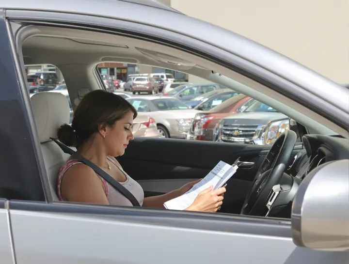Woman in her parked car checking a loan statement against her phone calculator