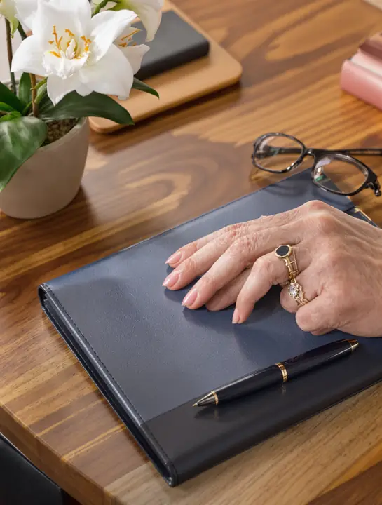 An older woman's hand resting on a navy portfolio with a pen beside it