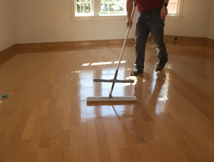 Refinisher spreading clear finish across a sanded floor with a T bar