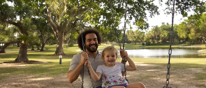 A father laughing with his daughter on a swing at a lakeside park