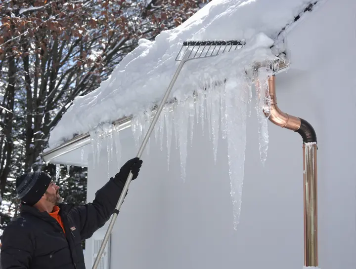 Homeowner raking snow off an old slate roof with ice dams on the gutter