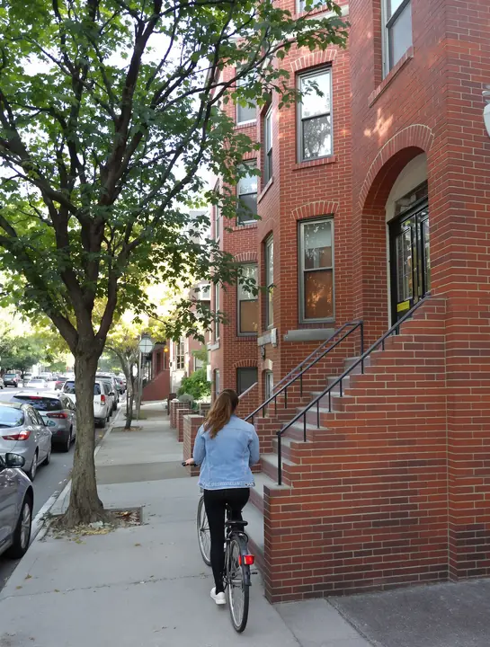 Woman carrying her road bike down a brick stoop on a leafy Astoria street