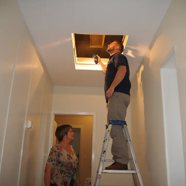 Estimator looking up through the attic hatch while the homeowner watches