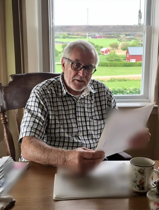 Retired farmer reading paperwork at his kitchen table with farmland through the window