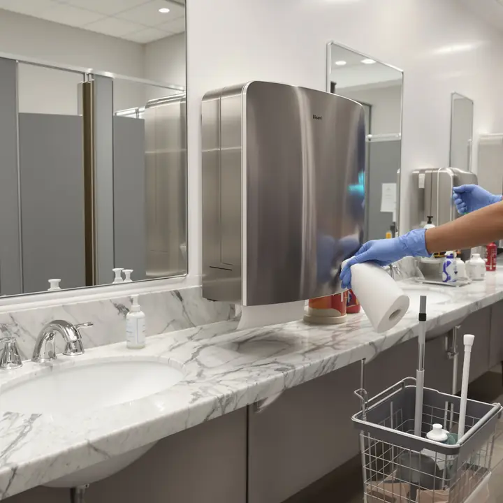 Gloved hands refilling a paper towel dispenser in an office restroom