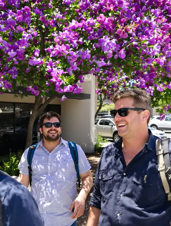 Two men in sunglasses laughing outside a building under purple jacaranda trees