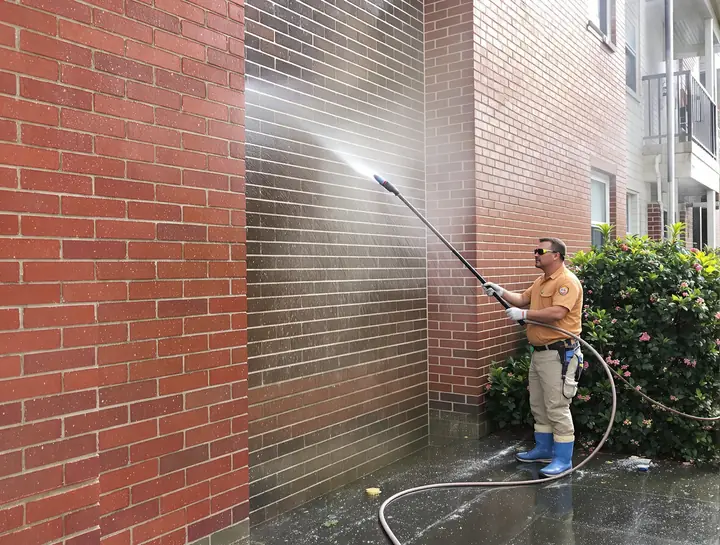 Worker pressure washing a brick apartment wall with a long wand