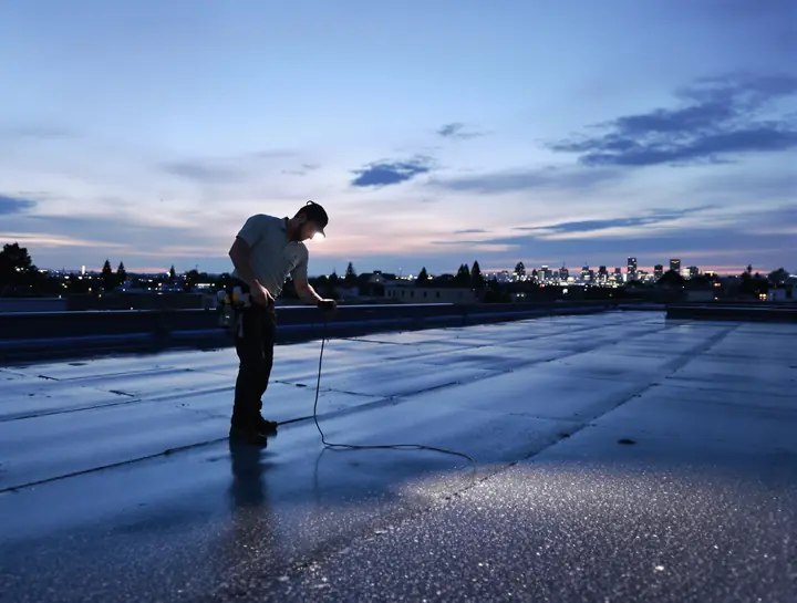 A roofing tech checking the membrane with a moisture meter at dusk
