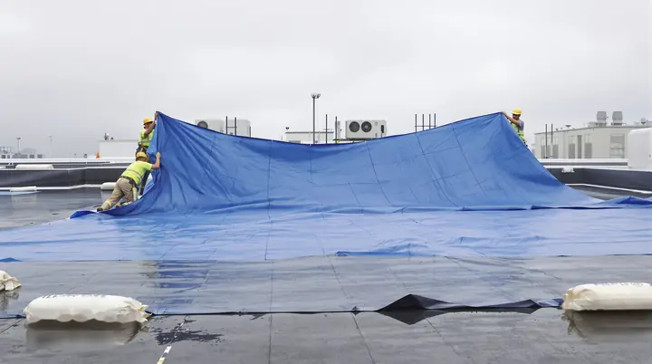A crew weighing down a tarp over a damaged roof section in the rain