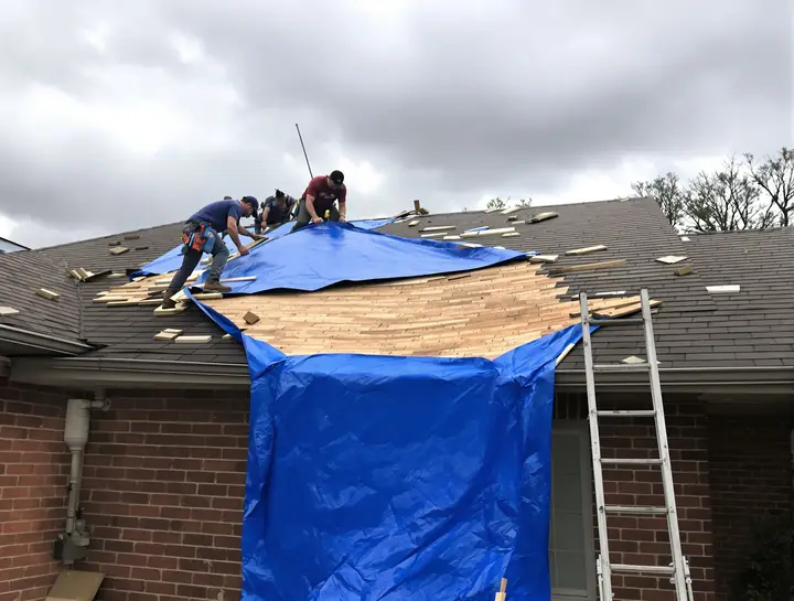 Two roofers nailing a blue tarp over wind torn shingles