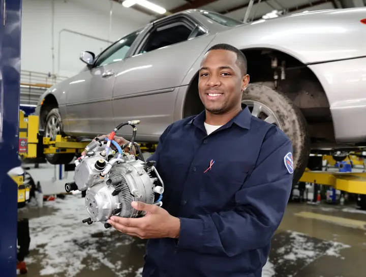 A mechanic holding a failed AC compressor under a lifted sedan